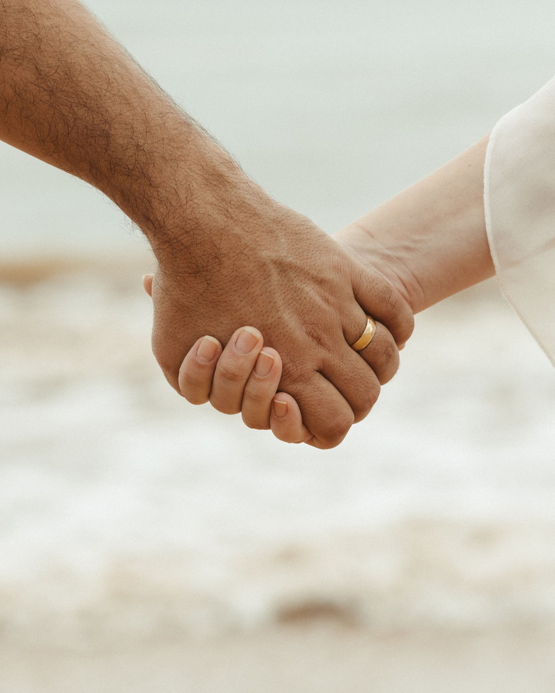 Close-up of two hands clasped together, a man's with a wedding ring and a woman's in a pale sleeve, against a soft blurred beach.